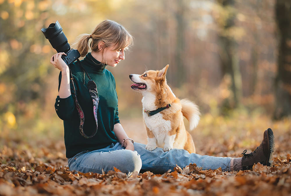 Female photographer with a dog sat down on the forest floor. She is holding a camera and the dog is looking at her