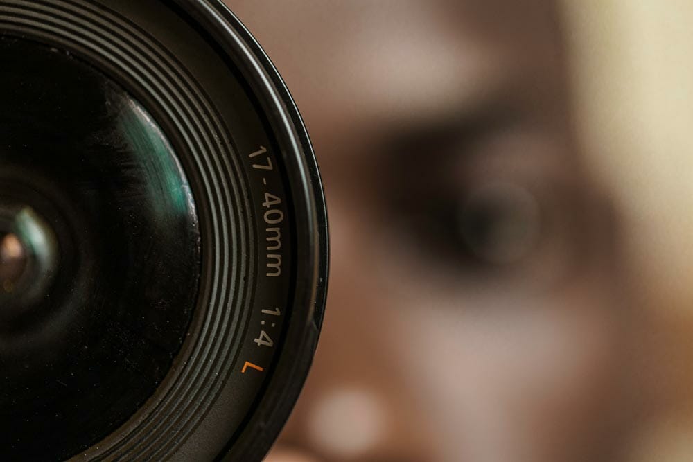 A close-up shot of a camera lens with a 17-40mm focal length. In the background, a young black man's head can be seen slightly out of focus